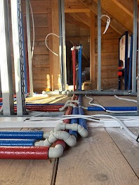 Instalacja wodna i kanalizacyjna w trakcie montażu w drewnianym domu. Widoczne rury w kolorze czerwonym i niebieskim oraz szara kanalizacja. Konstrukcja szkieletowa.