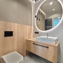 Nowoczesna łazienka z drewnianą zabudową, umywalką nablatową i okrągłym lustrem z podświetleniem LED oraz wiszącymi lampami.