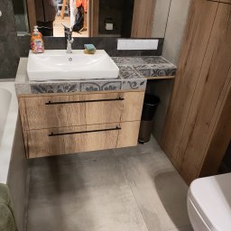 Sebastian Koła FURNIFUSION - Łazienka z drewnianą szafką pod umywalką z dwoma szufladami, blatem wykończonym dekoracyjnymi płytkami, oraz pasującą wysoką szafką obok, widoczna również toaleta i fragment wanny z ręcznikiem.
