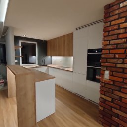 Sebastian Koła FURNIFUSION - Nowoczesna kuchnia z białymi i drewnianymi frontami, czarnym zlewem pod oknem i ceglanym filarem, oświetlenie LED pod szafkami.