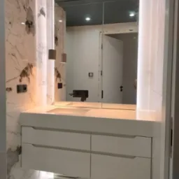 Nowoczesna łazienka z marmurowymi ścianami, białymi szafkami pod umywalką, lustrem z oświetleniem LED i wiszącymi lampami cylindrycznymi.