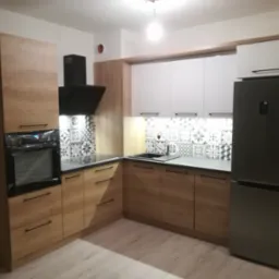 Nowoczesna kuchnia z drewnianymi frontami, czarnym okapem i srebrną lodówką, wykończona płytkami w geometryczne wzory. Piekarnik w folii ochronnej.