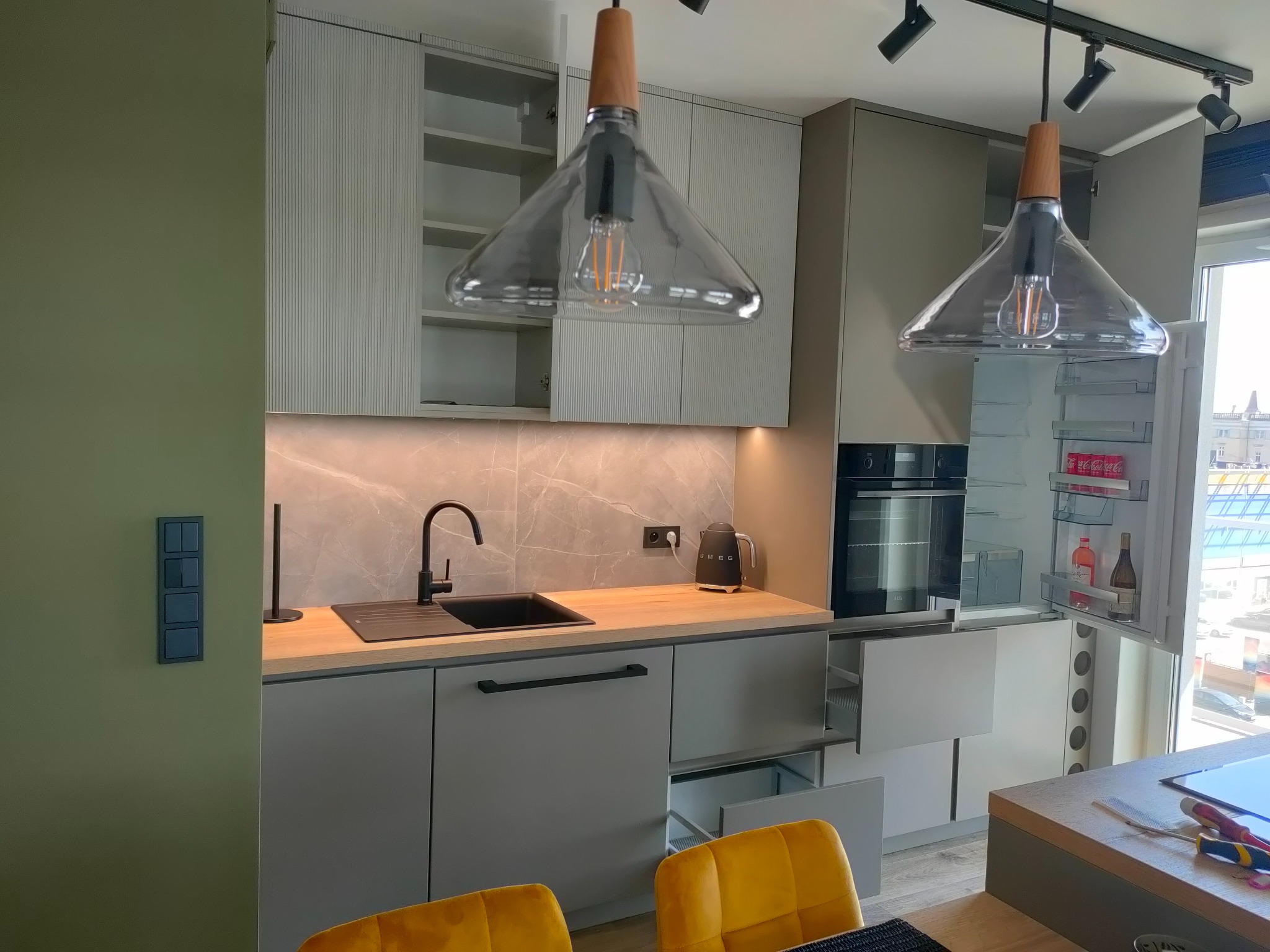 Nowoczesna kuchnia w szarościach z drewnianymi akcentami, oświetlona wiszącymi lampami z żarówkami Edisona i listwą reflektorów, z otwartą lodówką i wysuwanymi szufladami.