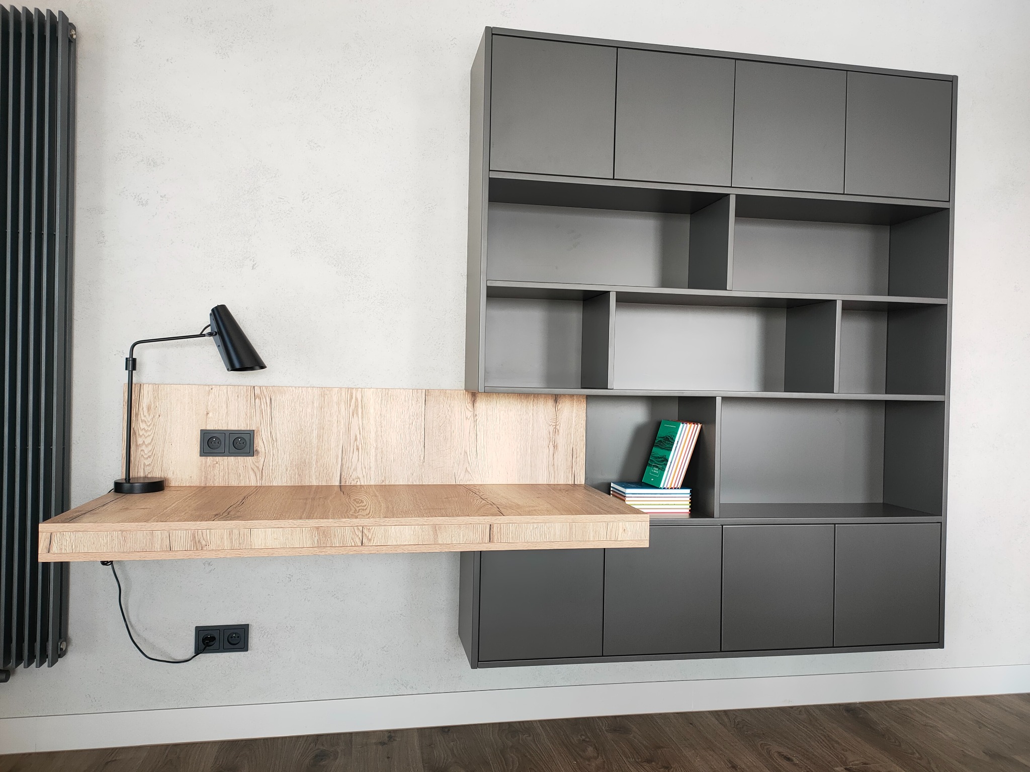 Szara zabudowa meblowa z biurkiem z drewnianym blatem, lampką i książkami na tle jasnej ściany z grzejnikiem.