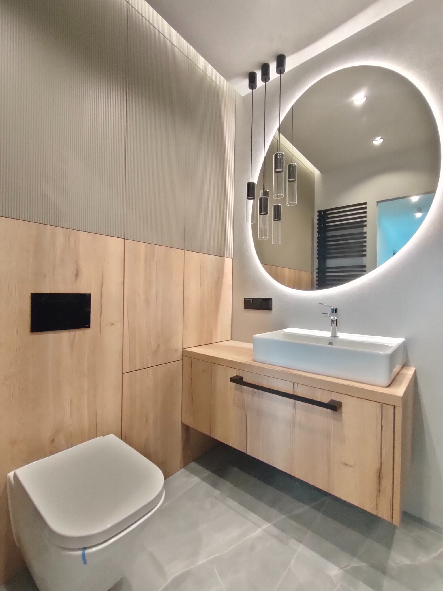 Nowoczesna łazienka z drewnianą zabudową, umywalką nablatową i okrągłym lustrem z podświetleniem LED oraz wiszącymi lampami.