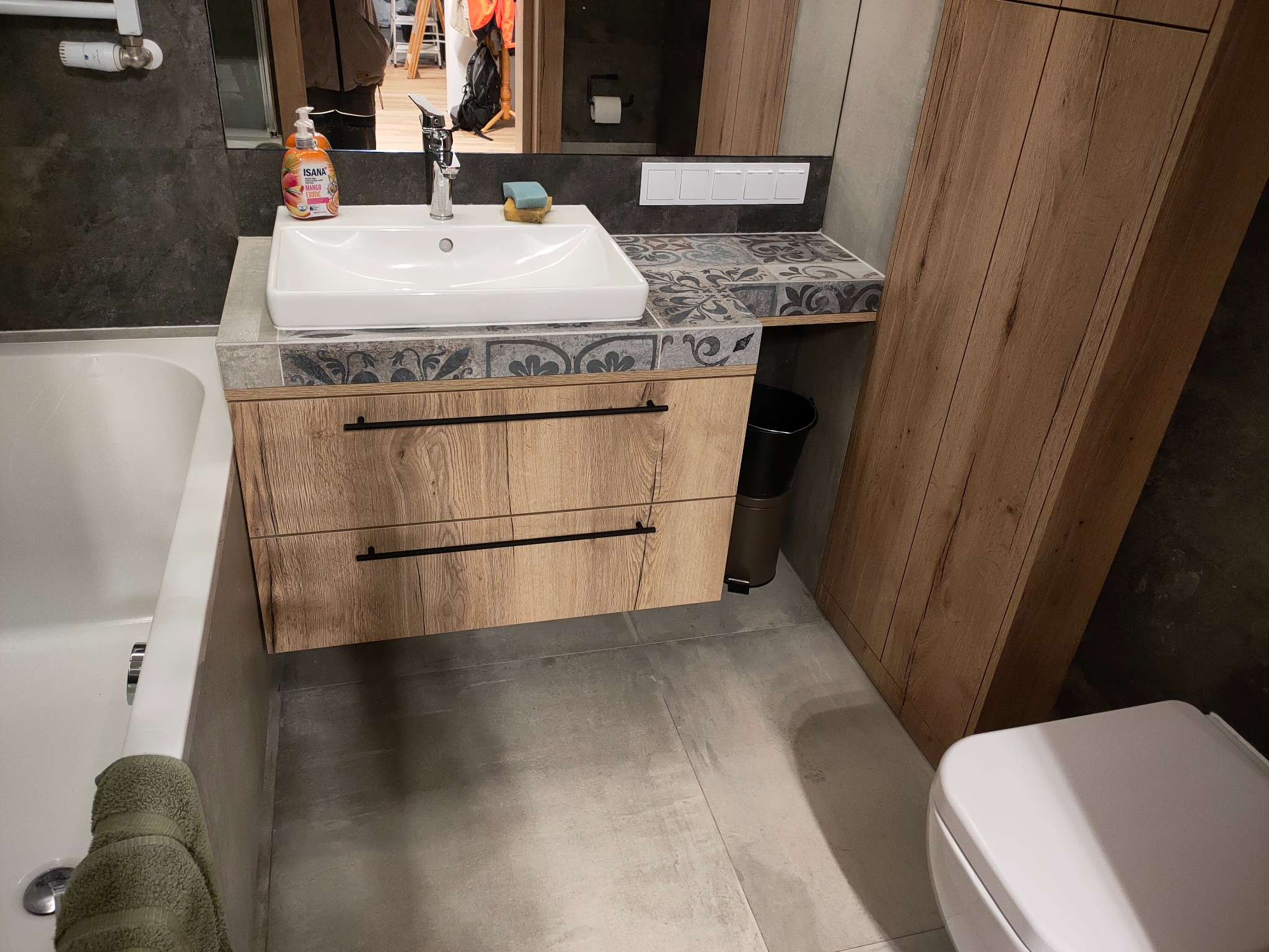 Łazienka z drewnianą szafką pod umywalką z dwoma szufladami, blatem wykończonym dekoracyjnymi płytkami, oraz pasującą wysoką szafką obok, widoczna również toaleta i fragment wanny z ręcznikiem.