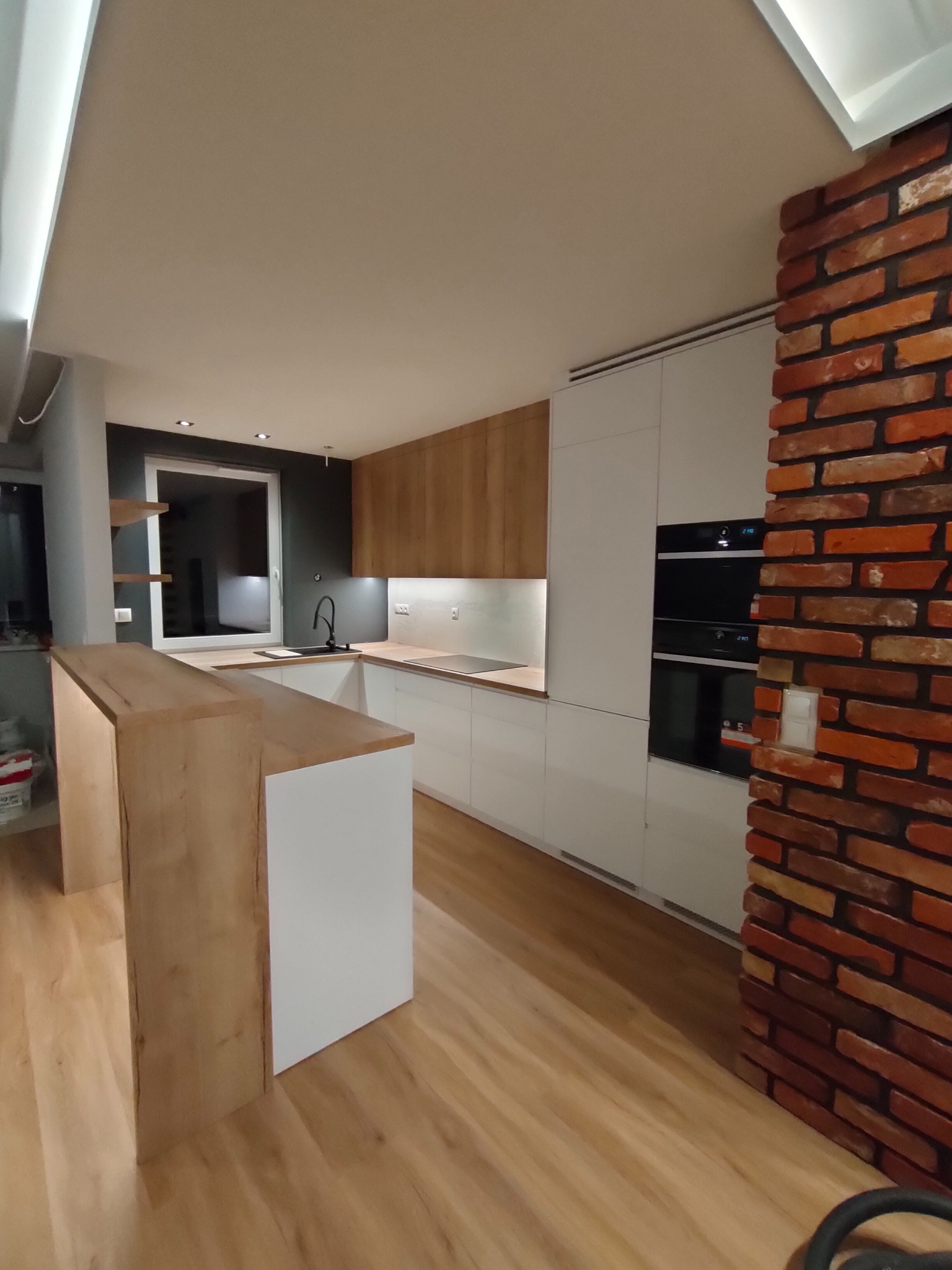 Nowoczesna kuchnia z białymi i drewnianymi frontami, czarnym zlewem pod oknem i ceglanym filarem, oświetlenie LED pod szafkami.