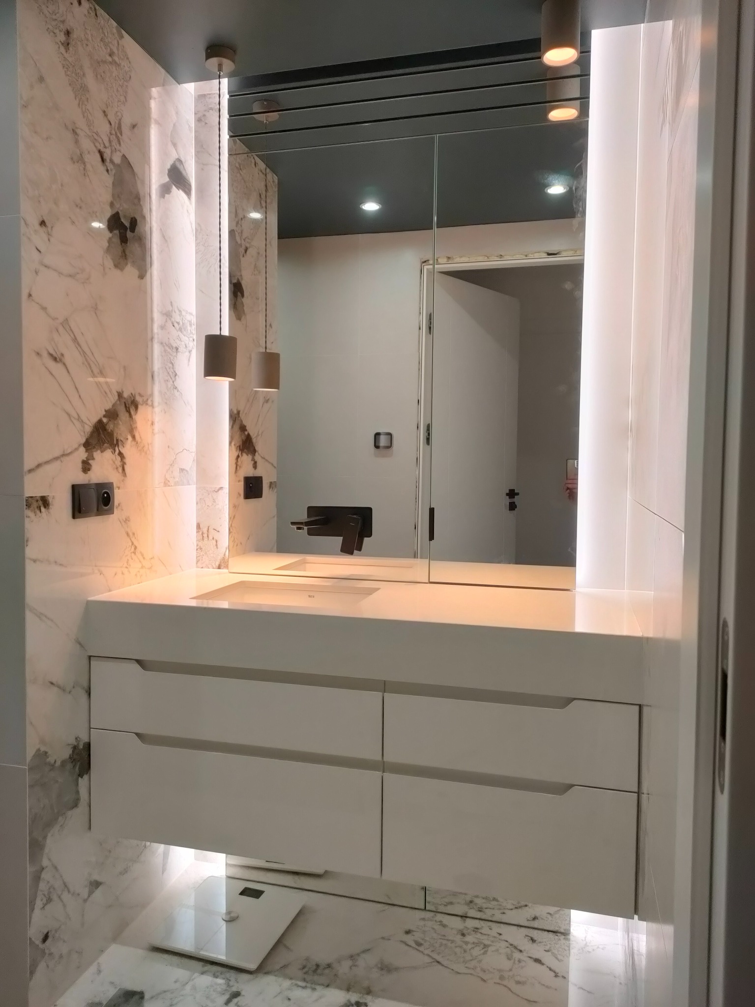 Nowoczesna łazienka z marmurowymi ścianami, białymi szafkami pod umywalką, lustrem z oświetleniem LED i wiszącymi lampami cylindrycznymi.
