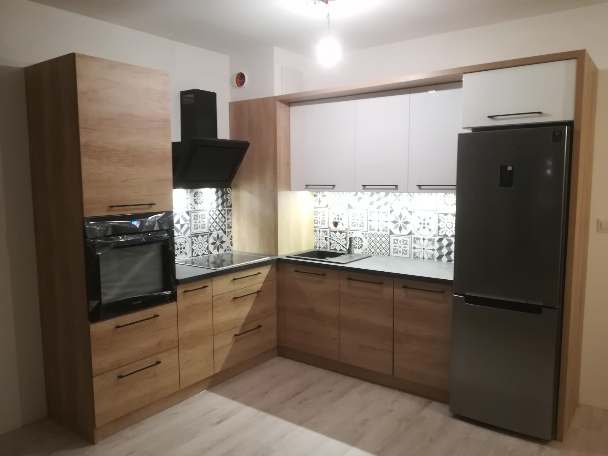 Nowoczesna kuchnia z drewnianymi frontami, czarnym okapem i srebrną lodówką, wykończona płytkami w geometryczne wzory. Piekarnik w folii ochronnej.