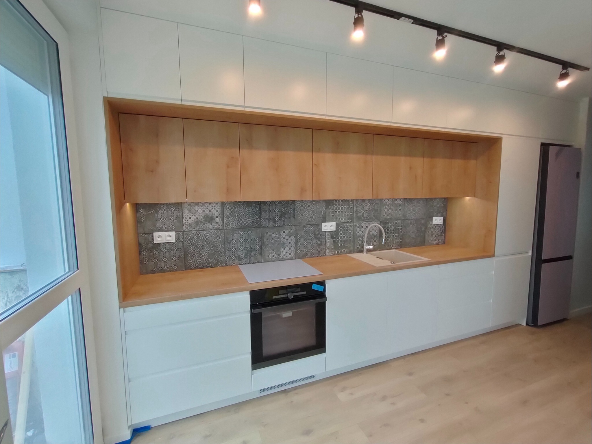 Nowoczesna kuchnia z białymi frontami szafek i drewnianymi akcentami, szarym backsplash z wzorem i oświetleniem punktowym na suficie. Widoczny piekarnik w zabudowie i lodówka.