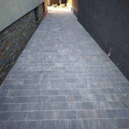 Big Myjka - Ułożona szara kostka brukowa tworząca ścieżkę między budynkami, widok z perspektywy pieszego, z elementami dekoracyjnymi na ścianie po lewej stronie.