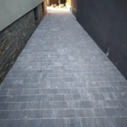 Ułożona szara kostka brukowa tworząca ścieżkę między budynkami, widok z perspektywy pieszego, z elementami dekoracyjnymi na ścianie po lewej stronie.