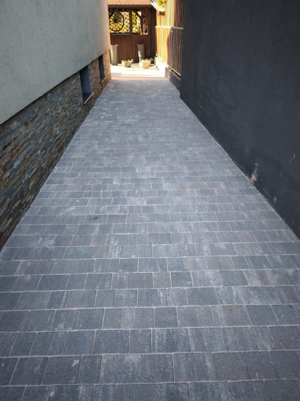 Ułożona szara kostka brukowa tworząca ścieżkę między budynkami, widok z perspektywy pieszego, z elementami dekoracyjnymi na ścianie po lewej stronie.