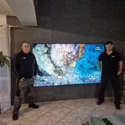 HORYZONT Montaż Serwis Anten TV Kraków - Dwóch mężczyzn pozuje na tle zamontowanego telewizora na ścianie z szarej płytki, widoczna wiązka lasera poziomującego. W tle zasłonięte okno i rośliny doniczkowe.