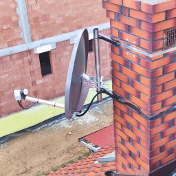 HORYZONT Montaż Serwis Anten TV Kraków - Antena satelitarna zamocowana do komina z cegły klinkierowej, z widocznym okablowaniem i elementami montażowymi, na tle budynku w trakcie budowy.