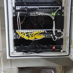 HORYZONT Montaż Serwis Anten TV Kraków - Szafa rack z zamontowanymi urządzeniami sieciowymi, w tym routerem Dahua, patch panelem z żółtymi kablami i switchem z zielonymi diodami, umieszczona w surowym pomieszczeniu z widocznymi kablami...