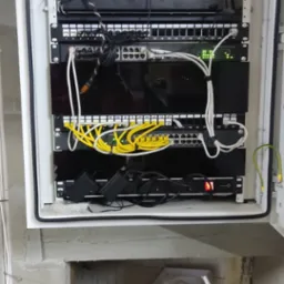 Szafa rack z zamontowanymi urządzeniami sieciowymi, w tym routerem Dahua, patch panelem z żółtymi kablami i switchem z zielonymi diodami, umieszczona w surowym pomieszczeniu z widocznymi kablami...