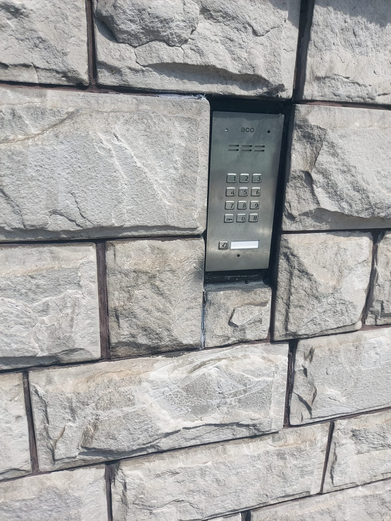 Zewnętrzny panel domofonu z klawiaturą numeryczną, zamontowany na elewacji z szarej cegły imitującej kamień.