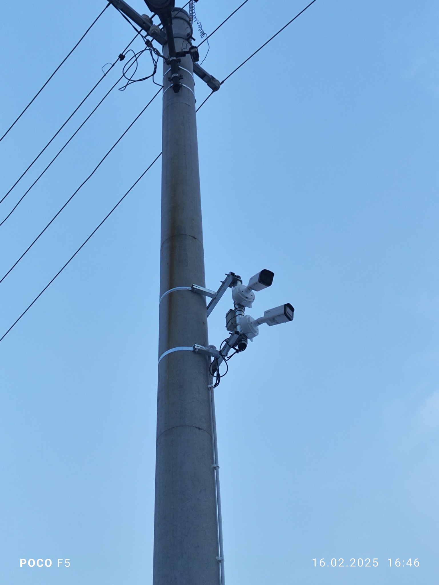 Dwie białe kamery monitoringu zamontowane na betonowym słupie energetycznym z widocznymi przewodami na tle błękitnego nieba.