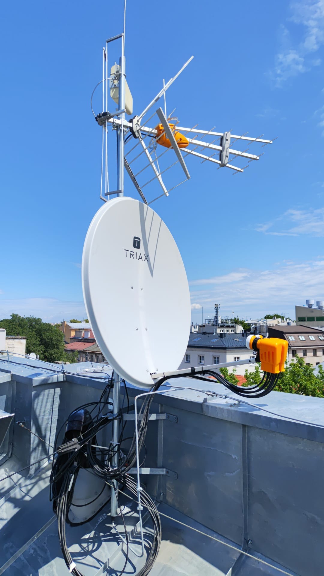 Zestaw antenowy na dachu budynku, antena satelitarna Triax, antena telewizyjna kierunkowa z pomarańczowym reflektorem, przewody antenowe. Widok na miasto w tle, błękitne niebo.