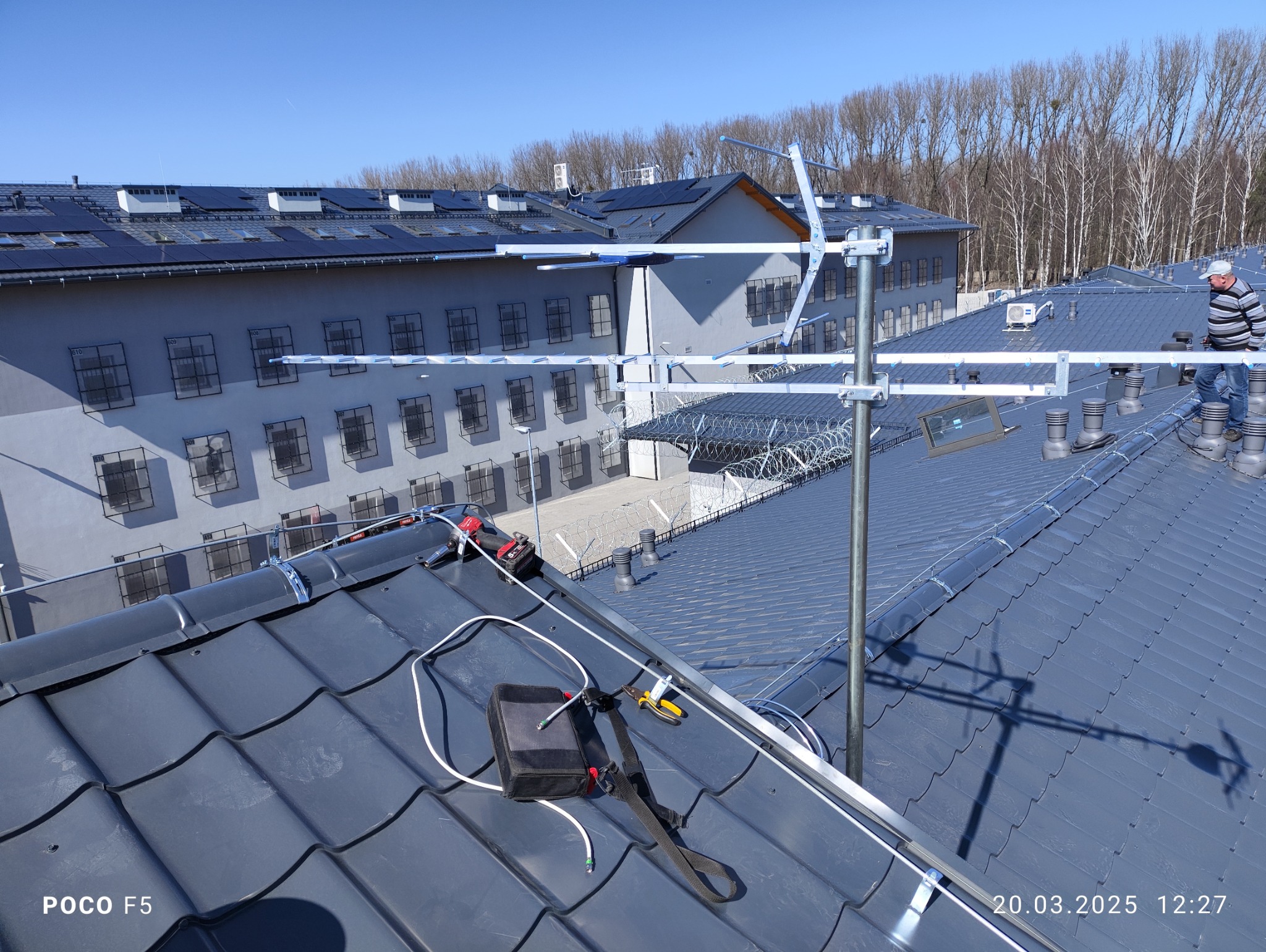 Instalacja anteny Yagi-Uda na dachu pokrytym ciemnoszarą dachówką ceramiczną; w tle budynek z kratami w oknach i drzewa bez liści.