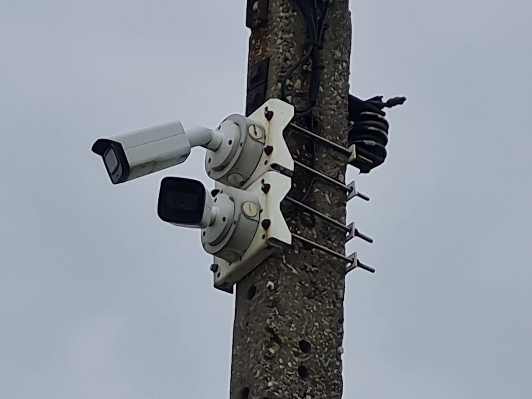 Dwie kamery monitoringu zamontowane na betonowym słupie, z widocznymi przewodami i metalowymi mocowaniami, na tle pochmurnego nieba.