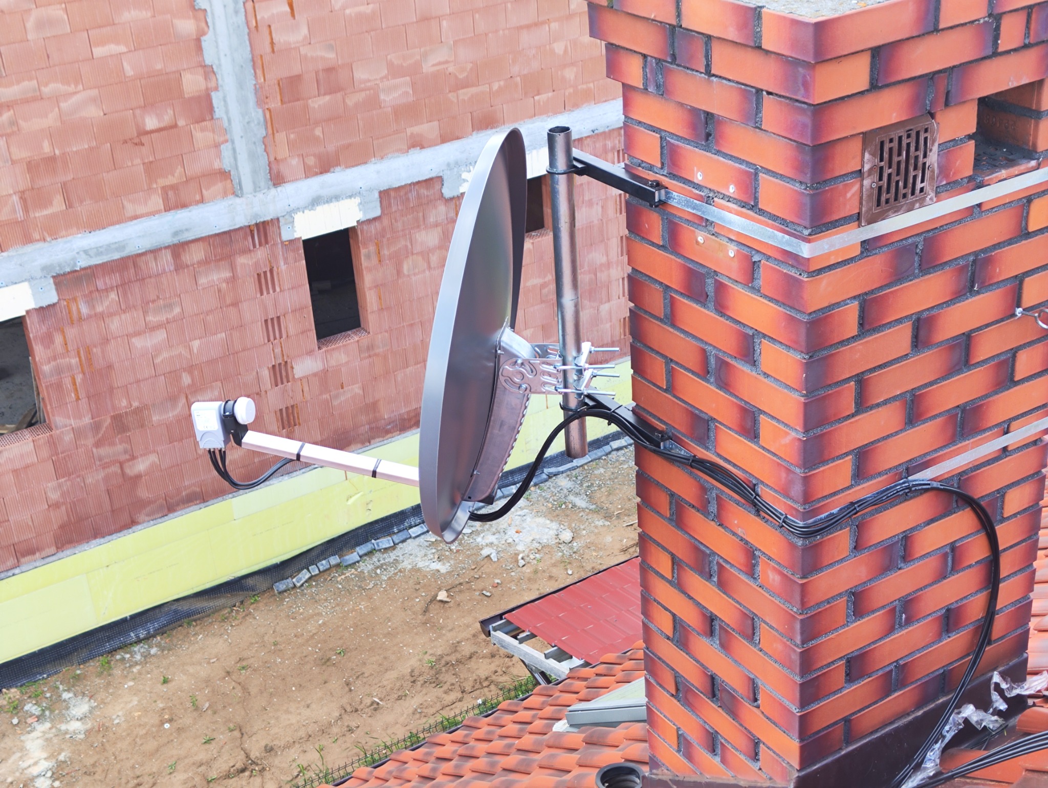 Antena satelitarna zamocowana do komina z cegły klinkierowej, z widocznym okablowaniem i elementami montażowymi, na tle budynku w trakcie budowy.