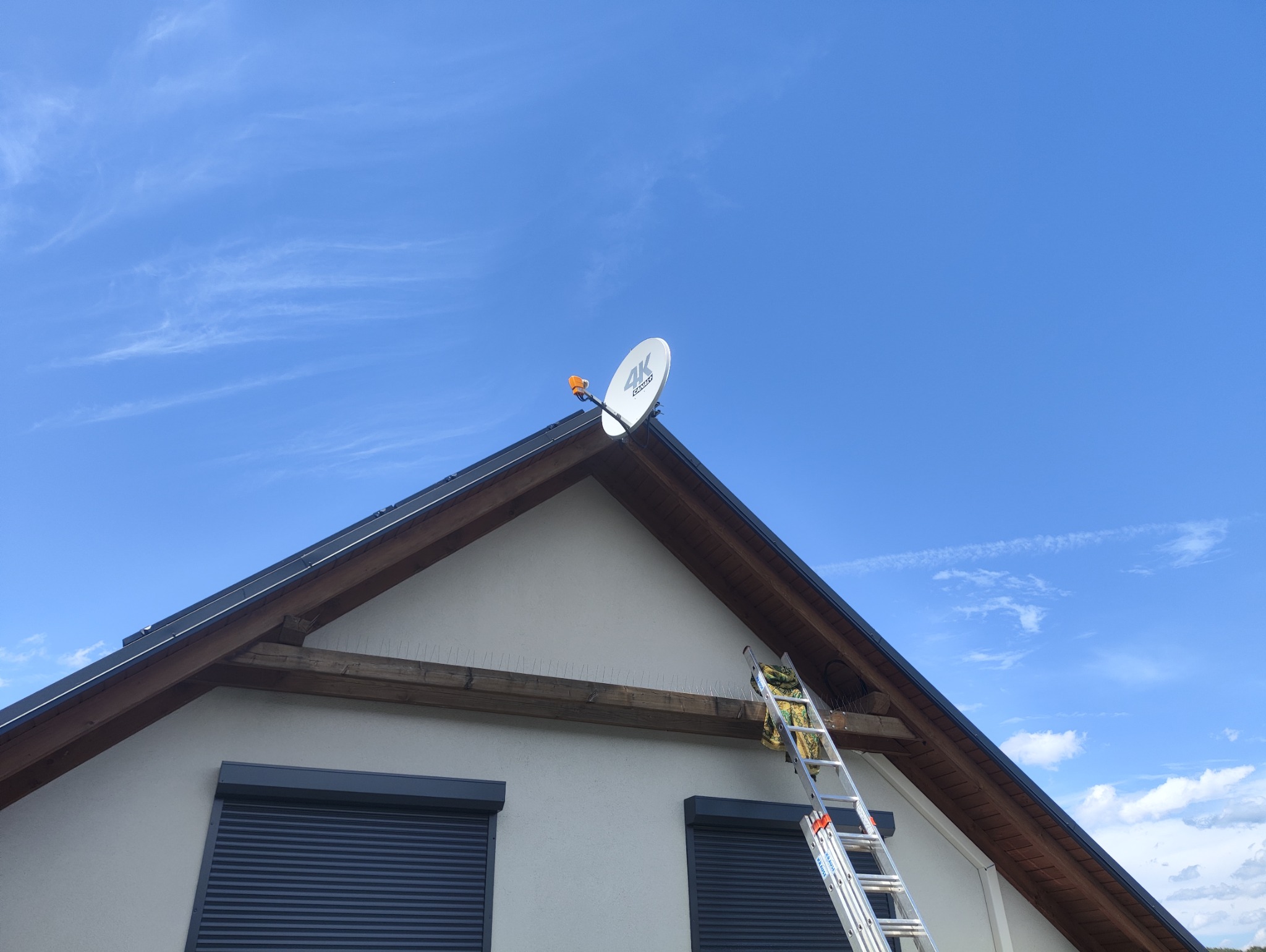 Biała antena satelitarna 4K zamontowana na dachu domu jednorodzinnego, z drabiny oparta o ścianę. Widoczne zabezpieczenia przeciw ptakom na belce pod dachem i błękitne niebo w tle.