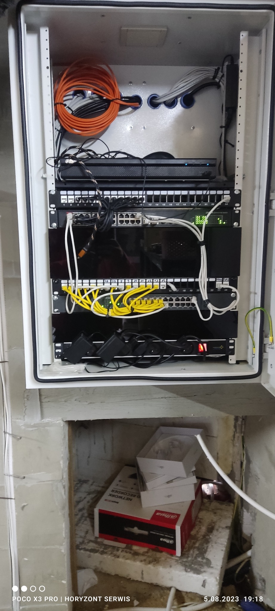 Szafa rack z zamontowanymi urządzeniami sieciowymi, w tym routerem Dahua, patch panelem z żółtymi kablami i switchem z zielonymi diodami, umieszczona w surowym pomieszczeniu z widocznymi kablami...