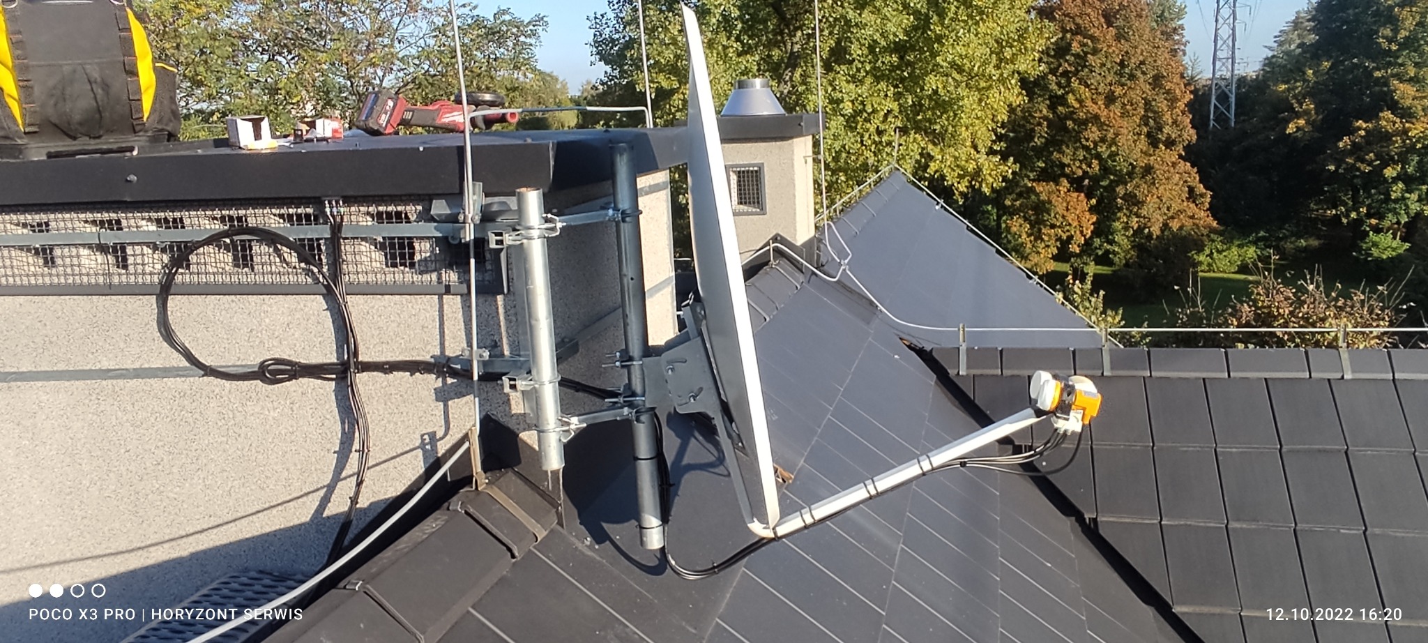 Antena satelitarna zamontowana na dachu budynku z widocznymi elementami mocującymi, kablem oraz narzędziami pracy w tle.