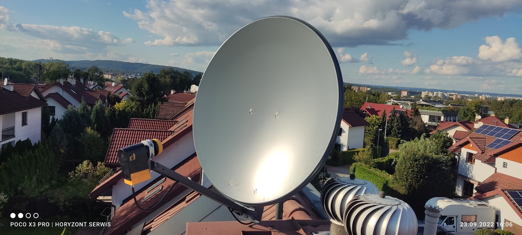 Antena satelitarna zamontowana na dachu domu z widokiem na okoliczną zabudowę i błękitne niebo z chmurami.