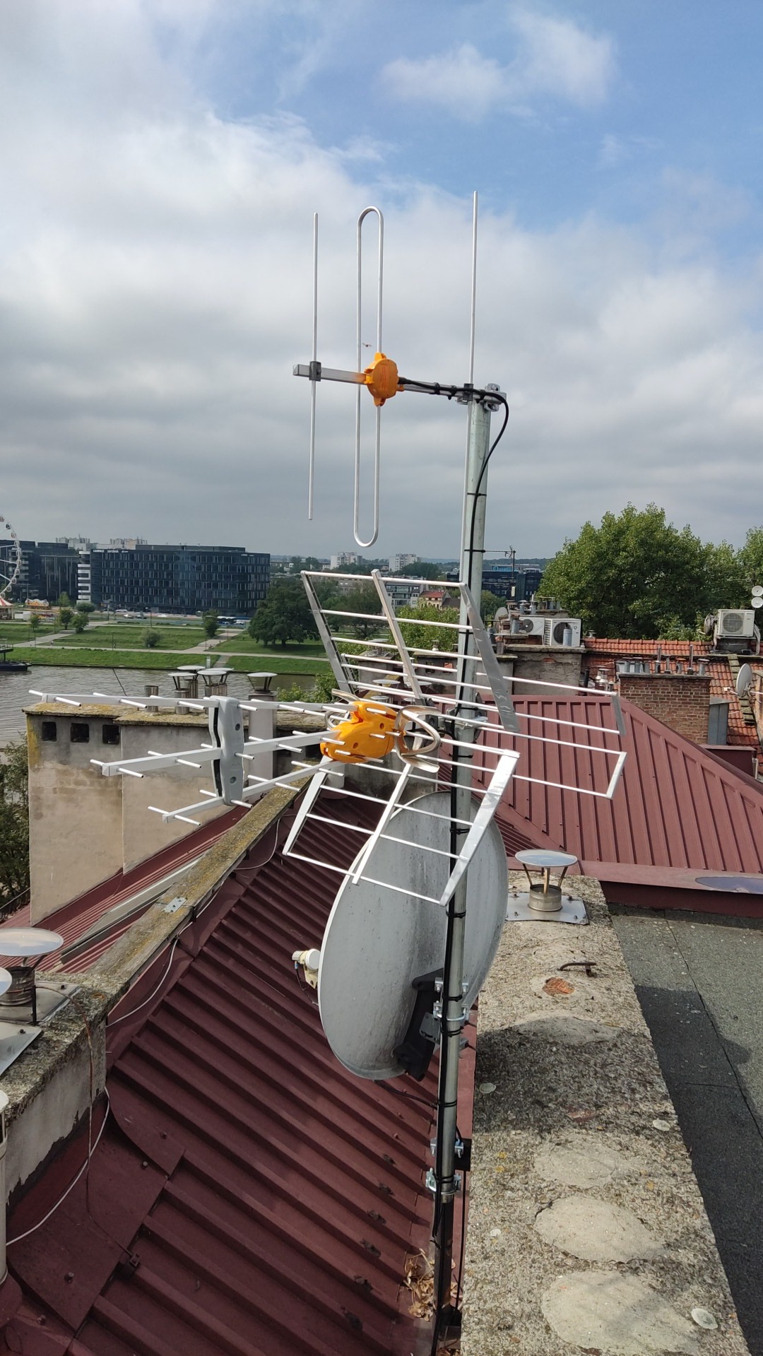 Zestaw antenowy UHF/VHF/SAT z pomarańczowymi elementami mocującymi, zainstalowany na dachu z czerwonej dachówki, w tle panorama miasta z rzeką i diabelskim młynem.
