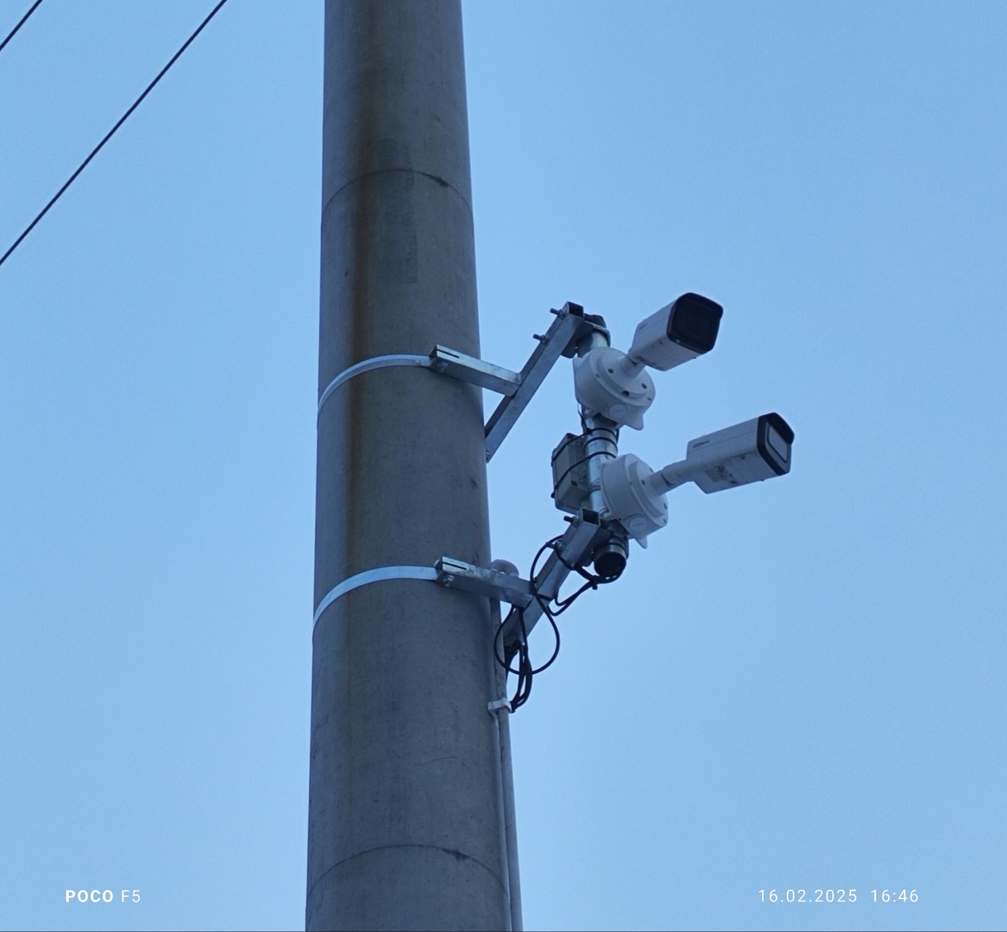 Dwie kamery monitoringu przymocowane do betonowego słupa za pomocą metalowych wsporników i obejm, widok z dołu na tle błękitnego nieba.