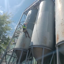 TYRMAR - Pracownik w uprzęży i kamizelce bezpieczeństwa czyści pod ciśnieniem duży, szary silos przemysłowy, wisząc na linach pod błękitnym niebem.