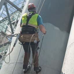 TYRMAR - Pracownik w uprzęży i kasku, z pistoletem natryskowym, maluje dużą cylindryczną konstrukcję przemysłową. W tle metalowe rusztowanie i błękitne niebo. Widoczne liny i sprzęt alpinistyczny.