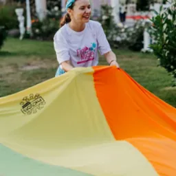 Uśmiechnięta kobieta w koszulce z logo 'Figle Migle' rozkłada kolorową chustę animacyjną na trawie podczas animacji na świeżym powietrzu.