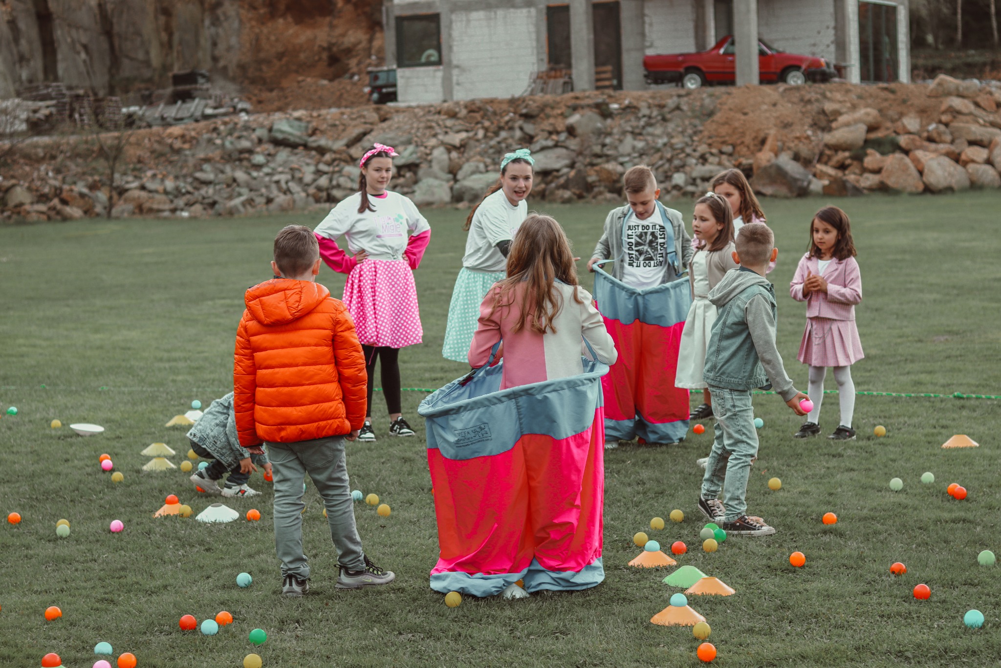 Grupa dzieci w różnym wieku uczestniczy w zabawie z kolorowymi workami i piłeczkami na trawie, w tle budynek w trakcie budowy i skalisty teren.