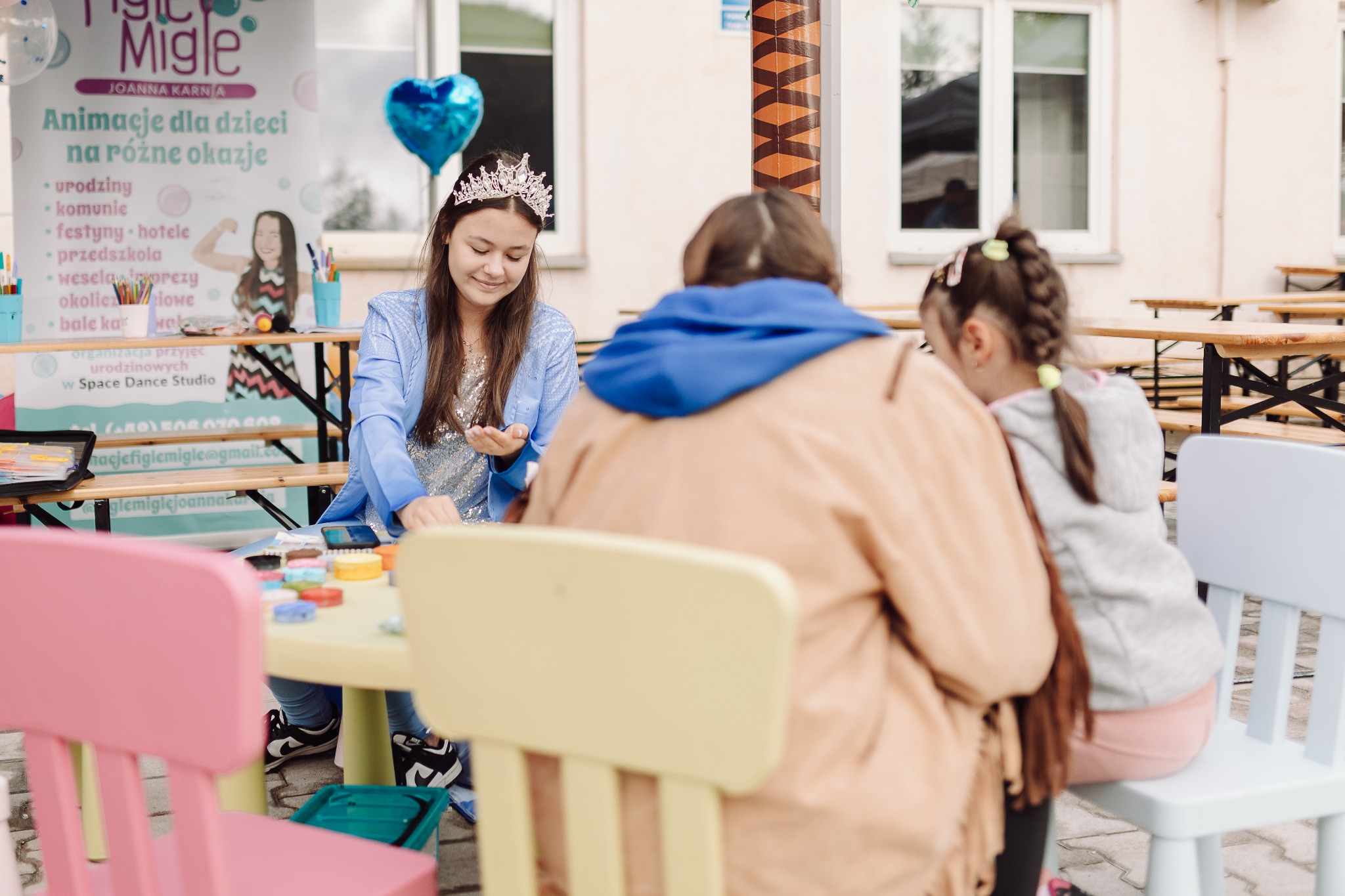 Dziewczynka w koronie i błyszczącej bluzce, siedząca przy stoliku z farbami do twarzy, w tle baner z napisem 'Animacje dla dzieci na różne okazje' i balon w kształcie serca.