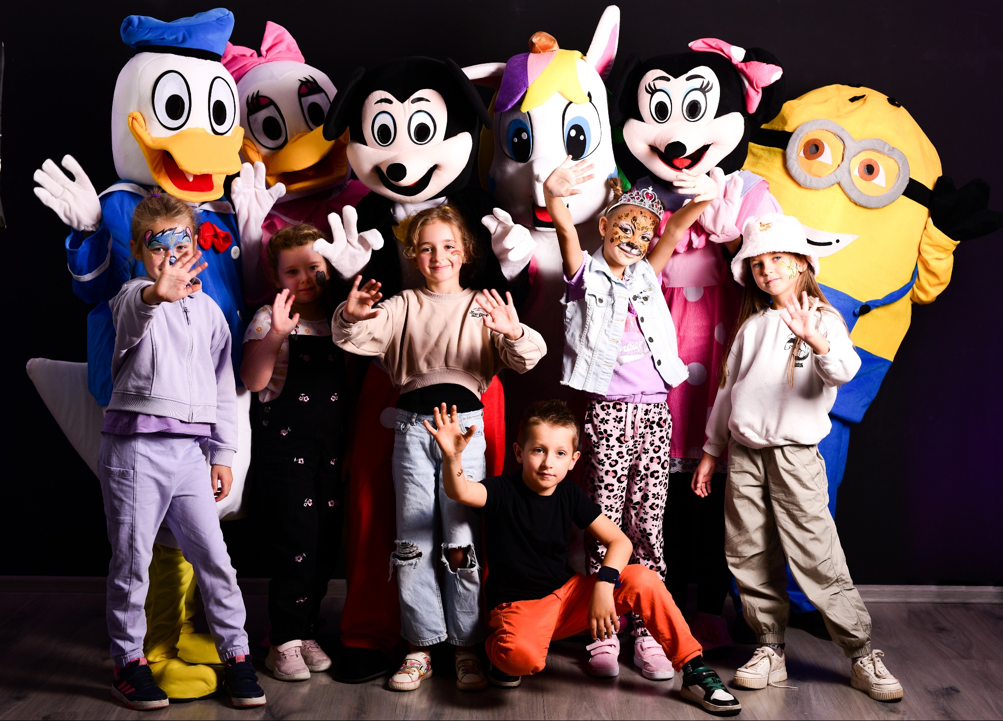 Grupa dzieci z pomalowanymi twarzami pozuje z animatorami przebranymi za postacie z bajek, w tym Kaczora Donalda, Myszki Miki i Minionka, na ciemnym tle.