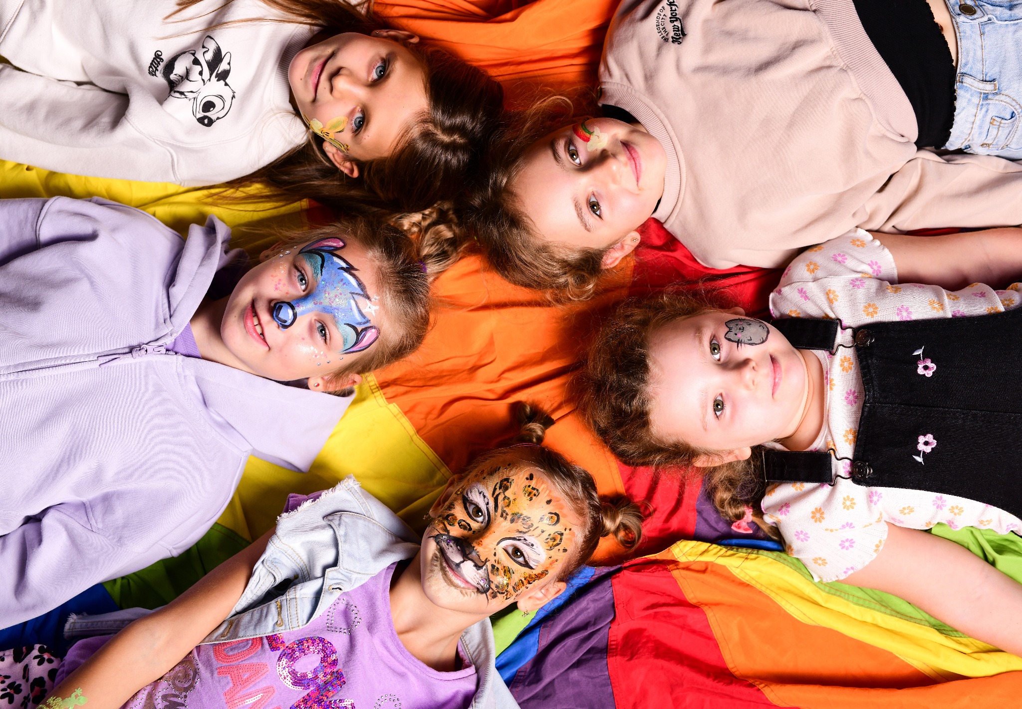 Pięć dziewczynek z pomalowanymi twarzami leży na kolorowej, tęczowej tkaninie, prezentując różne wzory makijażu: motyle, kotek, kwiaty i tygrys.
