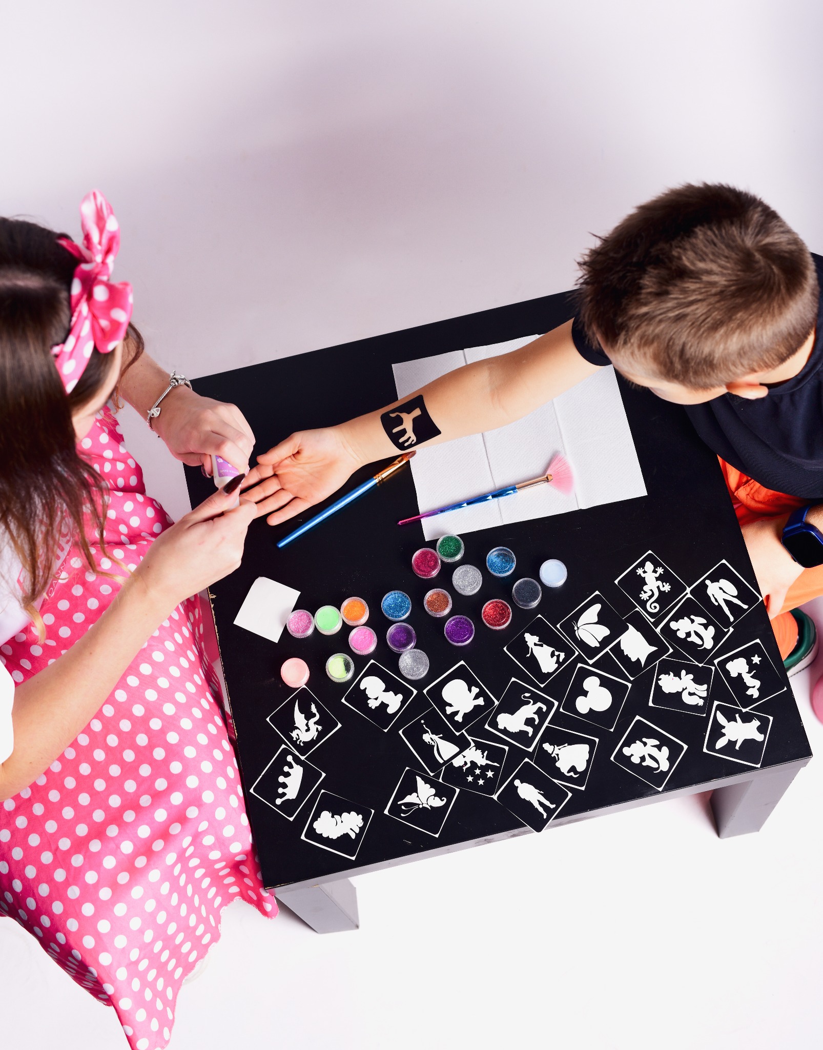 Nakładanie brokatowego tatuażu ze słonikiem na ramię dziecka przy użyciu szablonu; na stole rozłożone szablony z postaciami z bajek i brokaty.