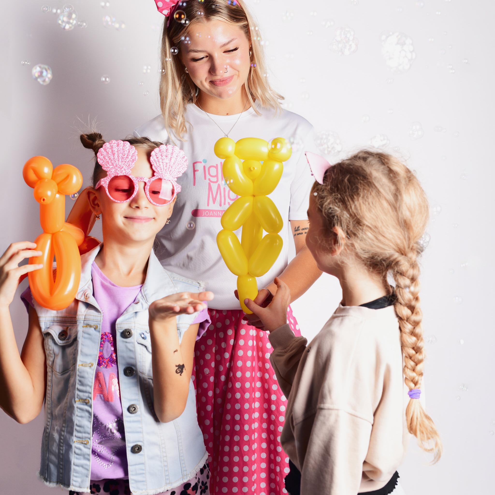 Kobieta w białej koszulce z różowym napisem i dwie dziewczynki bawiące się balonami w kształcie zwierząt i puszczające bańki mydlane na białym tle.