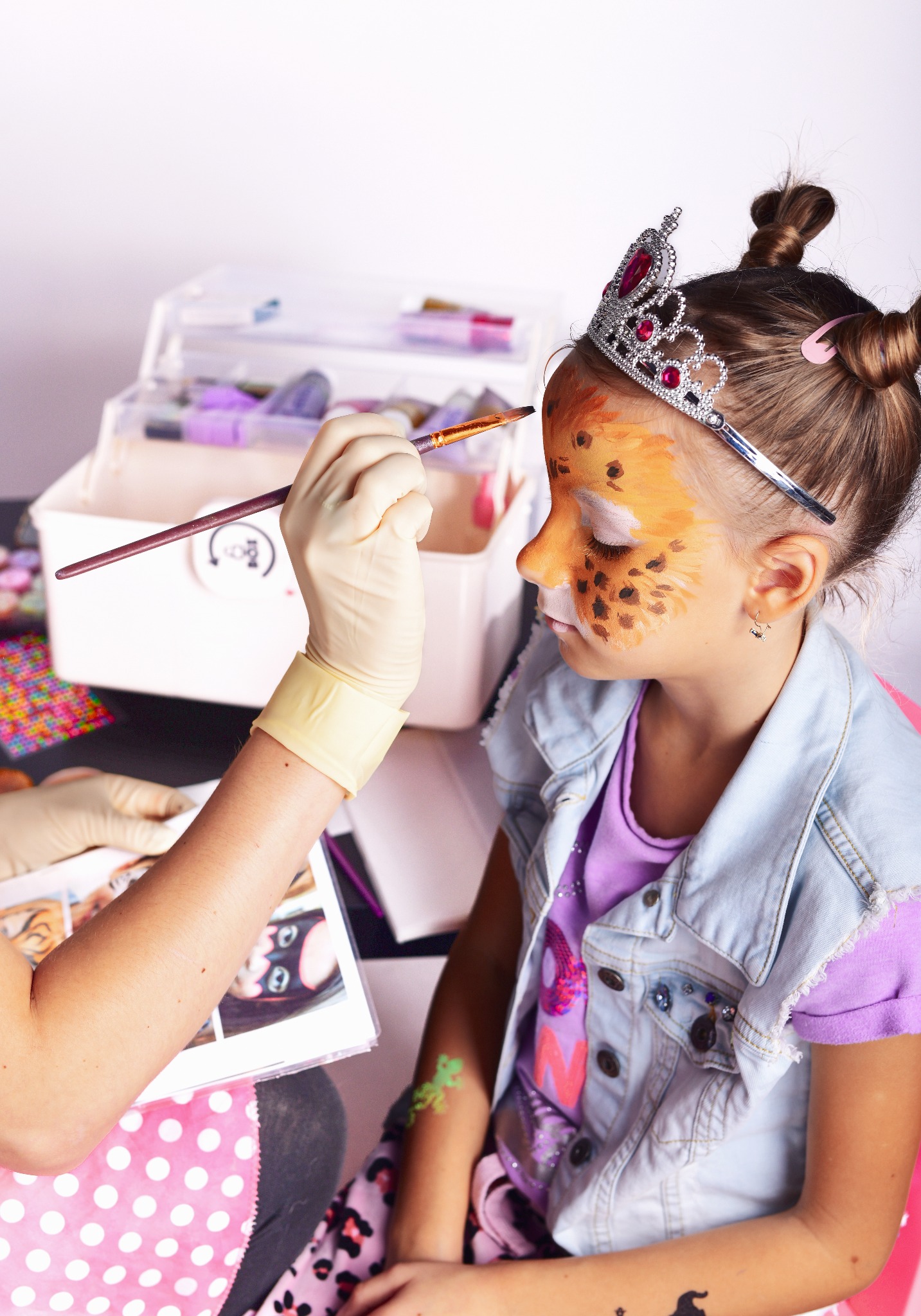 Dziewczynka z koroną na głowie, podczas malowania twarzy w motywy zwierzęce przez osobę w rękawiczkach, pędzel w dłoni, otoczenie stanowią akcesoria do malowania i wzornik.