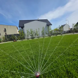 Intensywnie zielony trawnik nawadniany przez zraszacz rotacyjny, z widokiem na nowoczesne budynki i błękitne niebo z delikatnymi chmurami w tle.