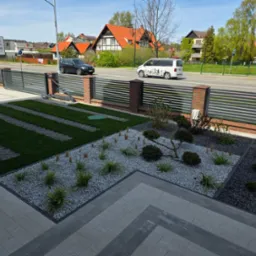 Nowoczesny ogród z geometrycznym układem trawnika i rabat wysypanych grysem, oddzielonych kostką. W tle posesja z ogrodzeniem i ulicą.