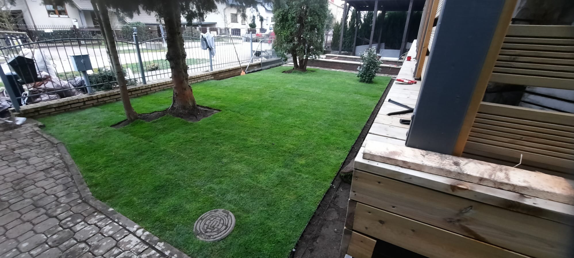 Świeżo ułożona trawa z rolki w ogrodzie z drzewami, widoczny fragment drewnianej konstrukcji tarasu i studzienka kanalizacyjna na trawniku.