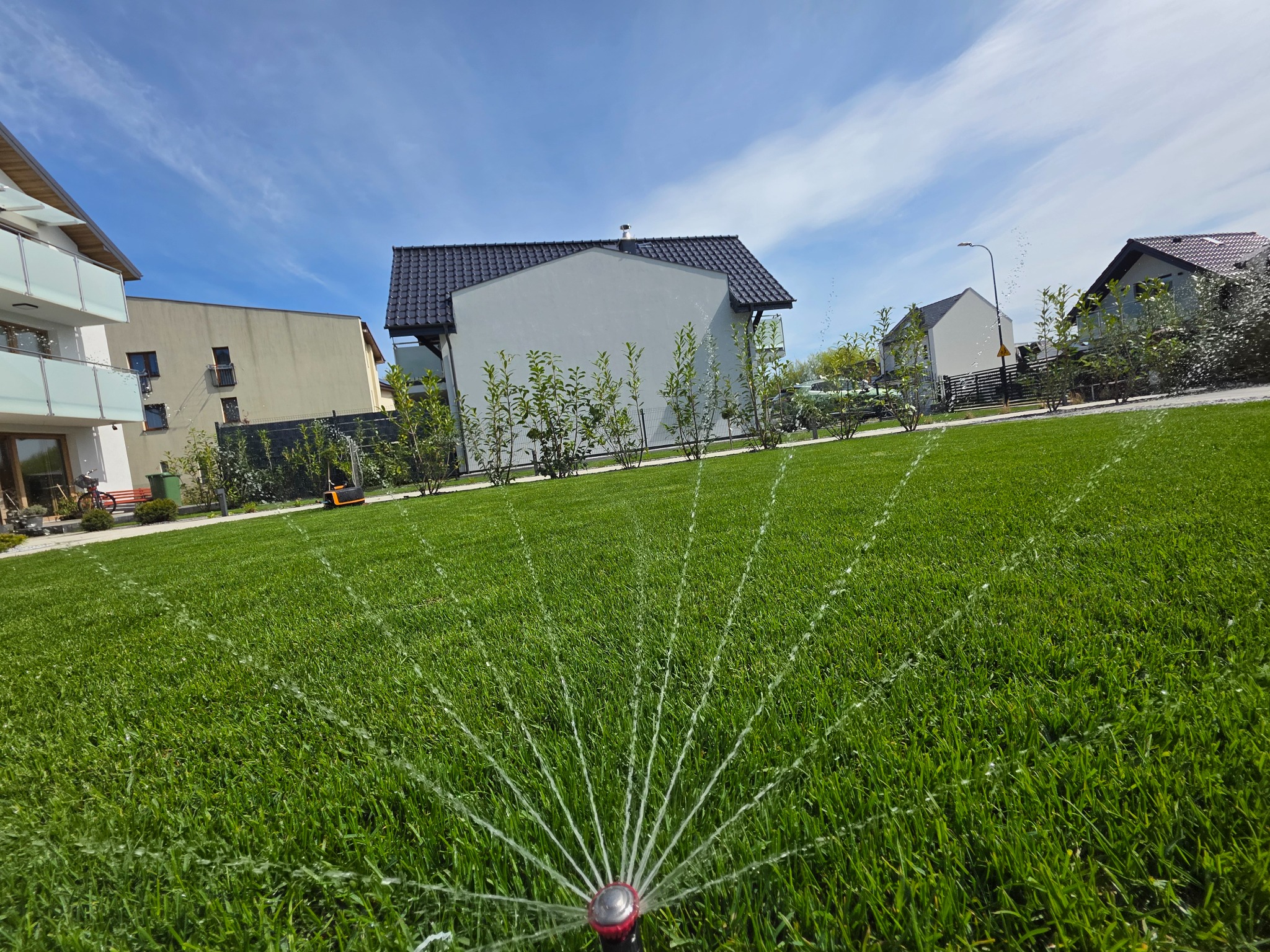 Intensywnie zielony trawnik nawadniany przez zraszacz rotacyjny, z widokiem na nowoczesne budynki i błękitne niebo z delikatnymi chmurami w tle.