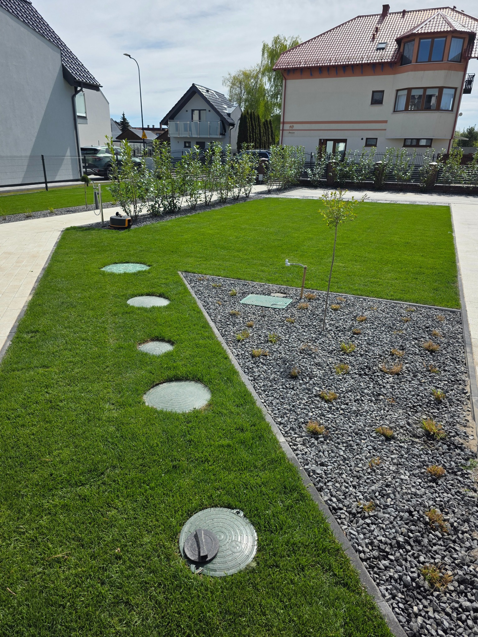 Zadbany trawnik z równo przystrzyżoną trawą, ozdobnym żwirem i młodymi krzewami na tle zabudowy jednorodzinnej w słoneczny dzień. Widoczne studzienki rewizyjne.