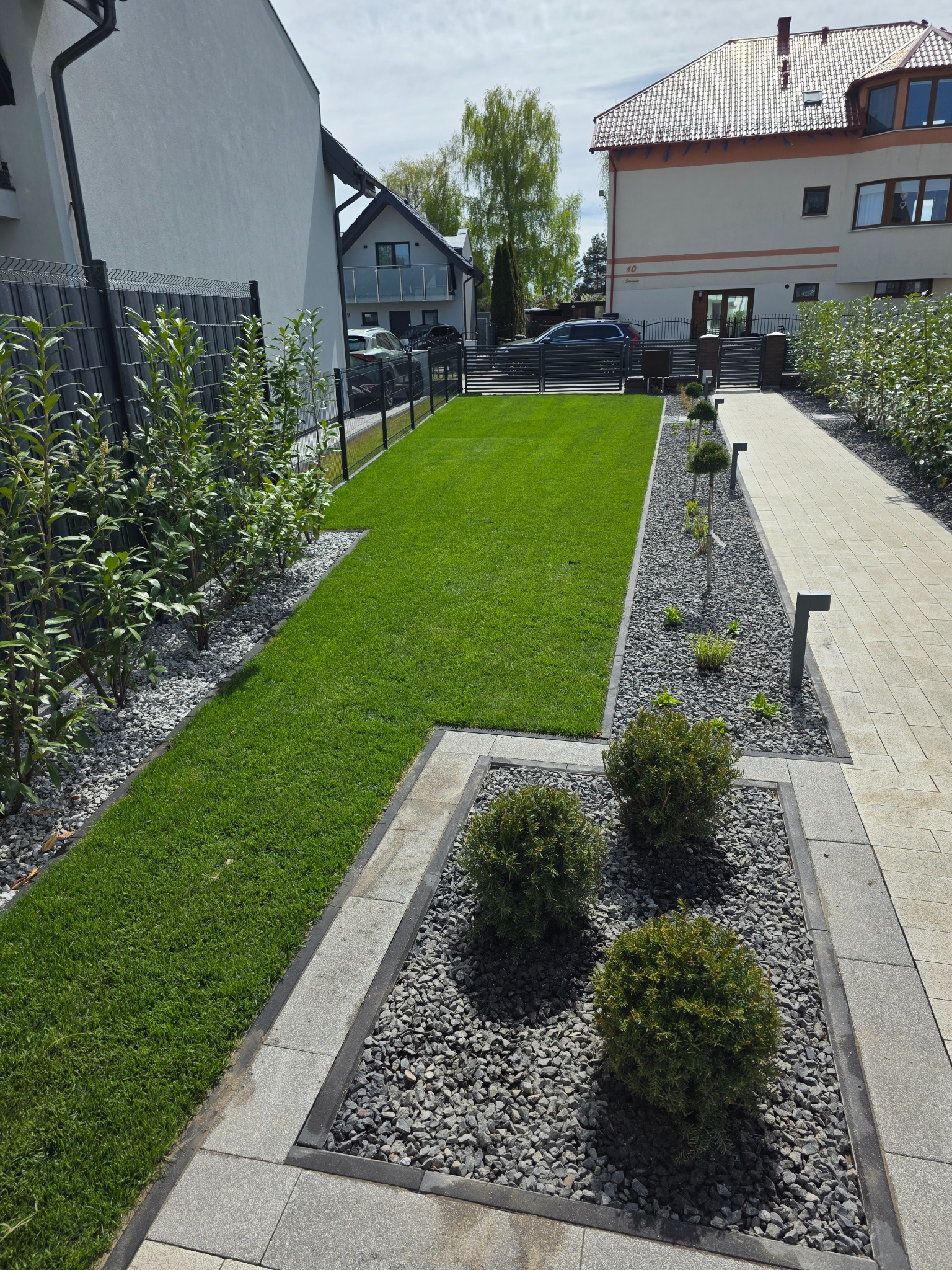 Zadbany ogród przydomowy z równo przystrzyżonym trawnikiem, ozdobnymi krzewami w szarych kamieniach i ścieżką z kostki, oświetlony lampami. W tle zabudowa jednorodzinna.