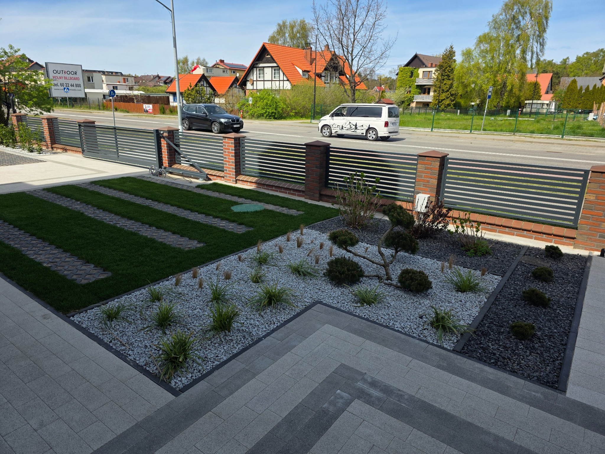 Nowoczesny ogród z geometrycznym układem trawnika i rabat wysypanych grysem, oddzielonych kostką. W tle posesja z ogrodzeniem i ulicą.
